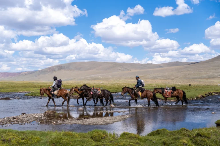 Twee ruiters steken met hun paarden een rivier over in een uitgestrekt bergachtig landschap.