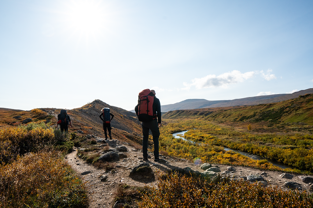 Twee wandelaars met grote rugzakken lopen over een stenig pad door een bergdal met herfstkleuren en een rivier.