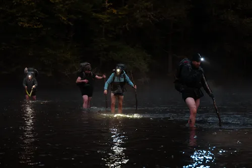 Vier wandelaars met hoofdlampen en rugzakken steken 's nachts een donkere rivier over.