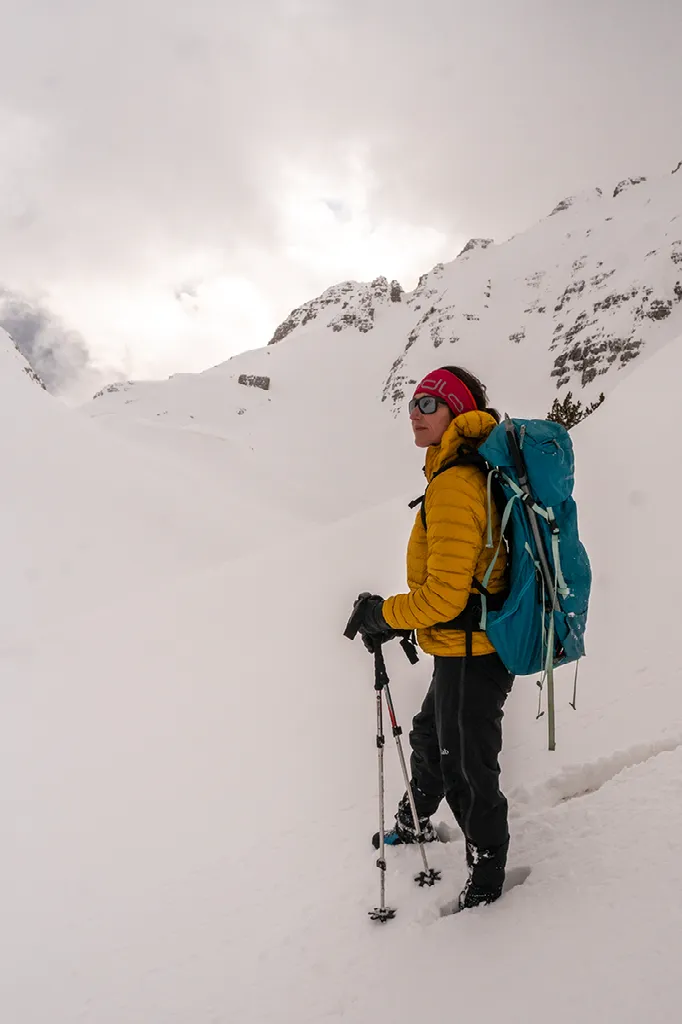Een wandelaar met een gele jas, blauwe rugzak en wandelstokken staat in diepe sneeuw met besneeuwde bergen op de achtergrond.
