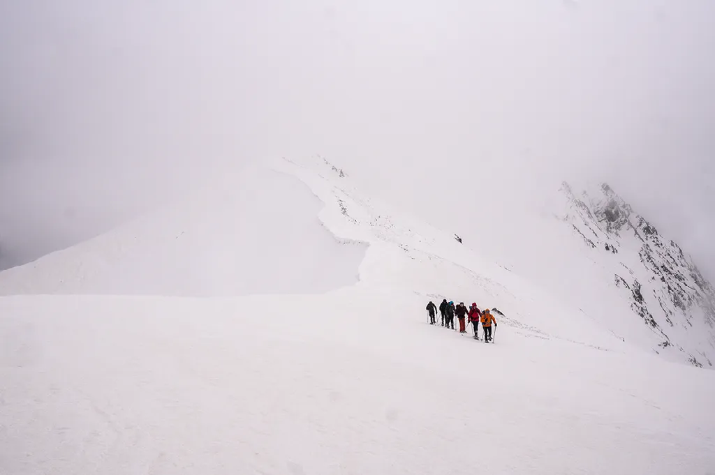 Een groep van zes wandelaars loopt achter elkaar over een steile, besneeuwde berghelling in dichte mist.