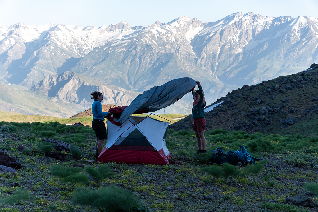 Twee mensen zetten een tent op in een berglandschap met besneeuwde toppen op de achtergrond