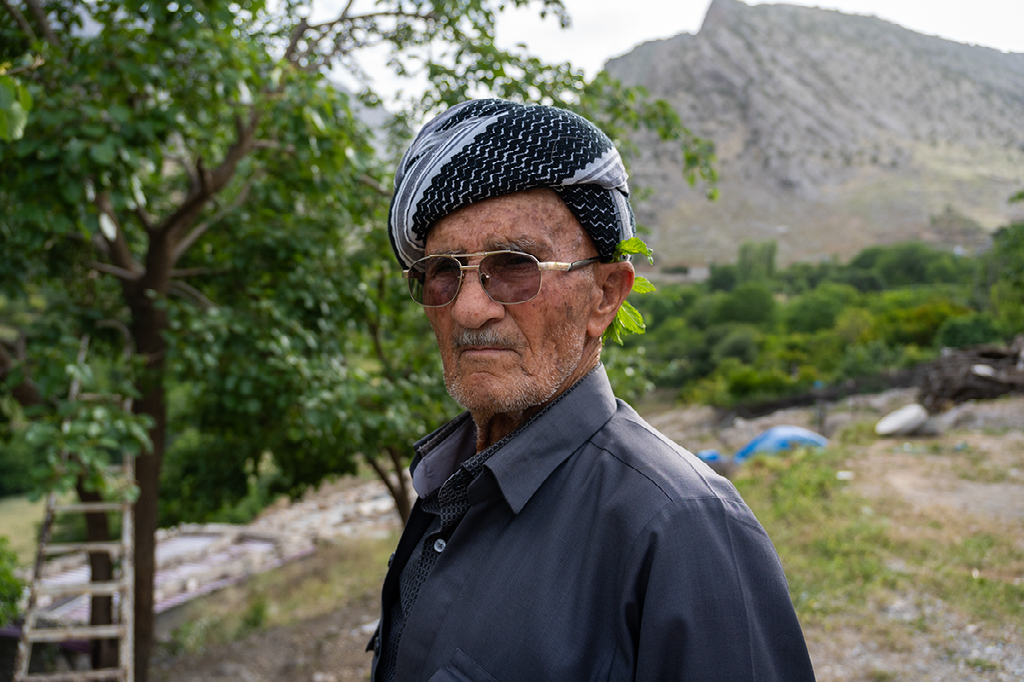 portret van een oudere Koerdische man met zonnebril en traditionele hoofddoek in een bergdorp met groene omgeving