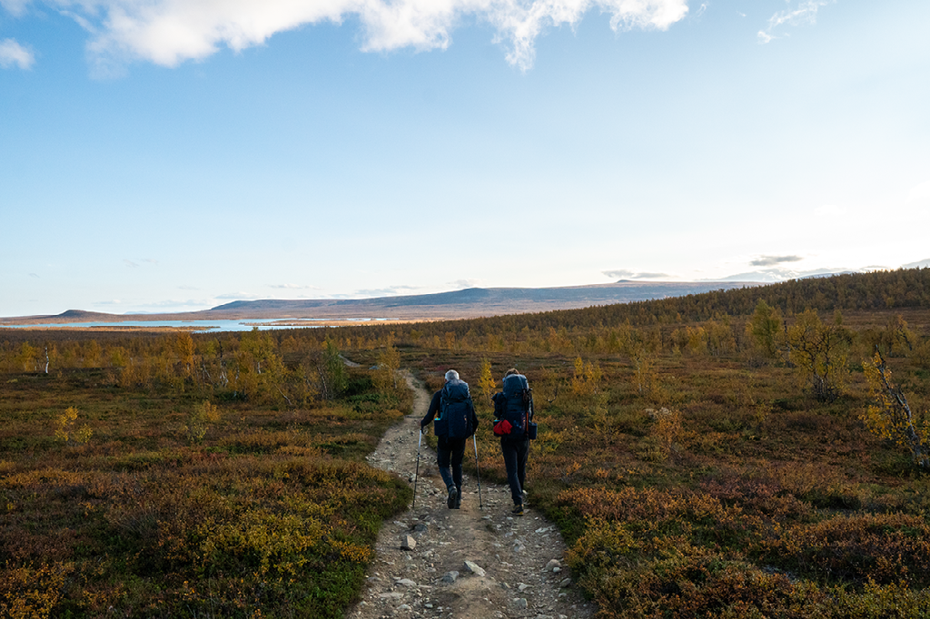 Twee wandelaars met rugzakken lopen over een zandpad door een glooiend toendra-landschap met herfstkleuren onder een helderblauwe lucht.