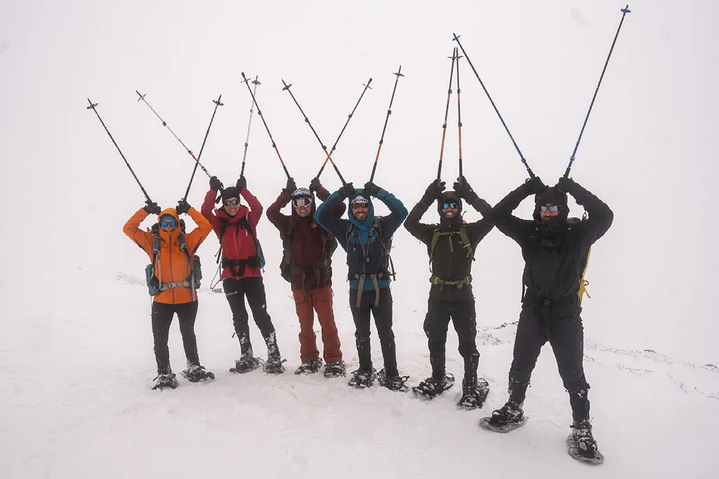 Een groep van zes wandelaars in winterkleding met sneeuwschoenen en stokken juicht met de armen omhoog op een besneeuwde helling.