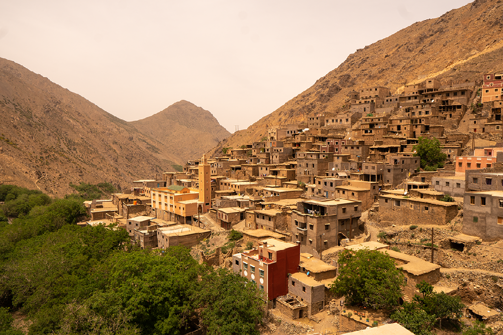 Een dichtbebouwd Berberdorp met traditionele stenen huizen en een minaret ligt tegen een steile, droge berghelling.