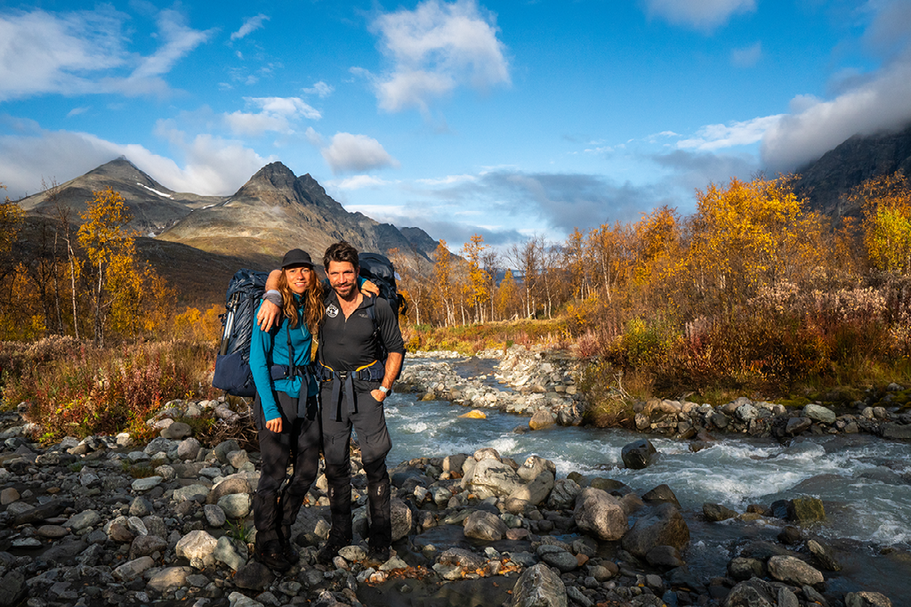 Twee lachende wandelaars met rugzakken staan op een rotsachtige oever bij een snelstromende rivier, met op de achtergrond bergen en herfstkleurige bomen.