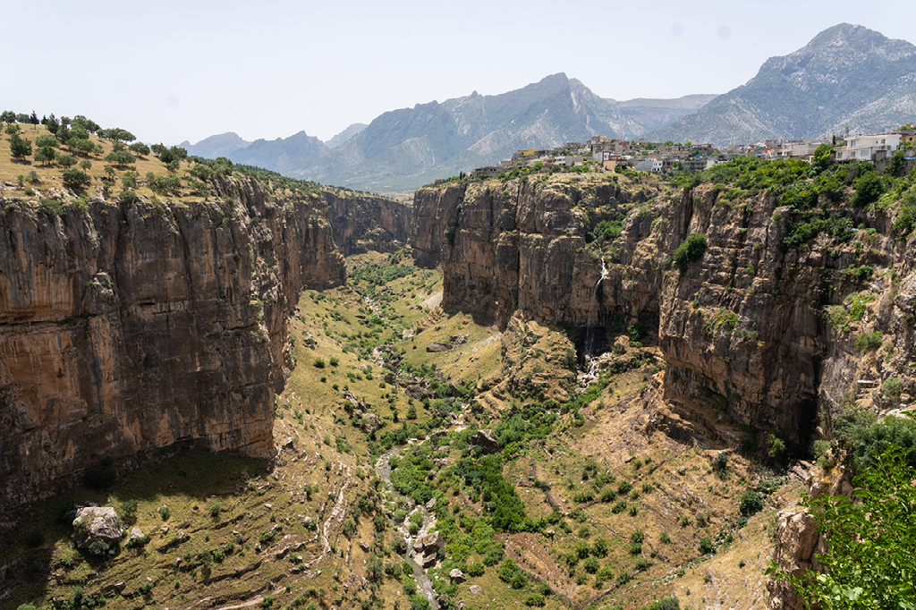 Een diepe groene kloof omringd door steile rotswanden met op de achtergrond een bergachtig landschap en een dorpje boven op de rand