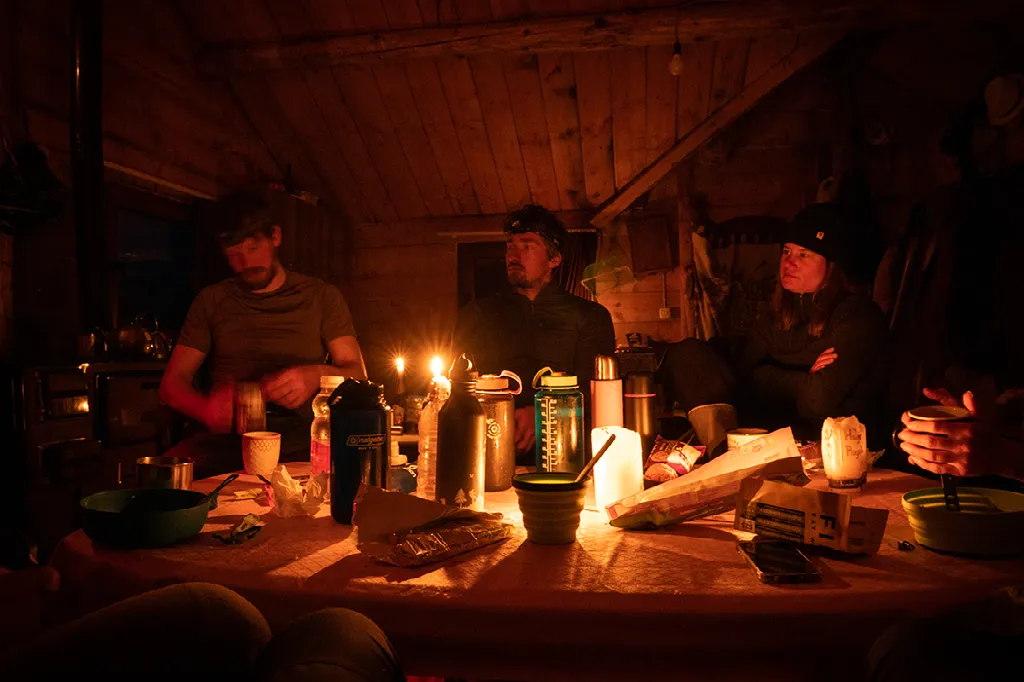 Drie wandelaars zitten 's avonds bij kaarslicht aan een houten tafel in een hut met mokken en eten.