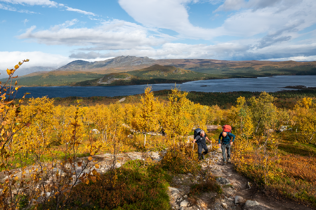 Twee wandelaars lopen over een pad door gele en oranje herfstbomen met uitzicht op een groot meer en bergen op de achtergrond.
