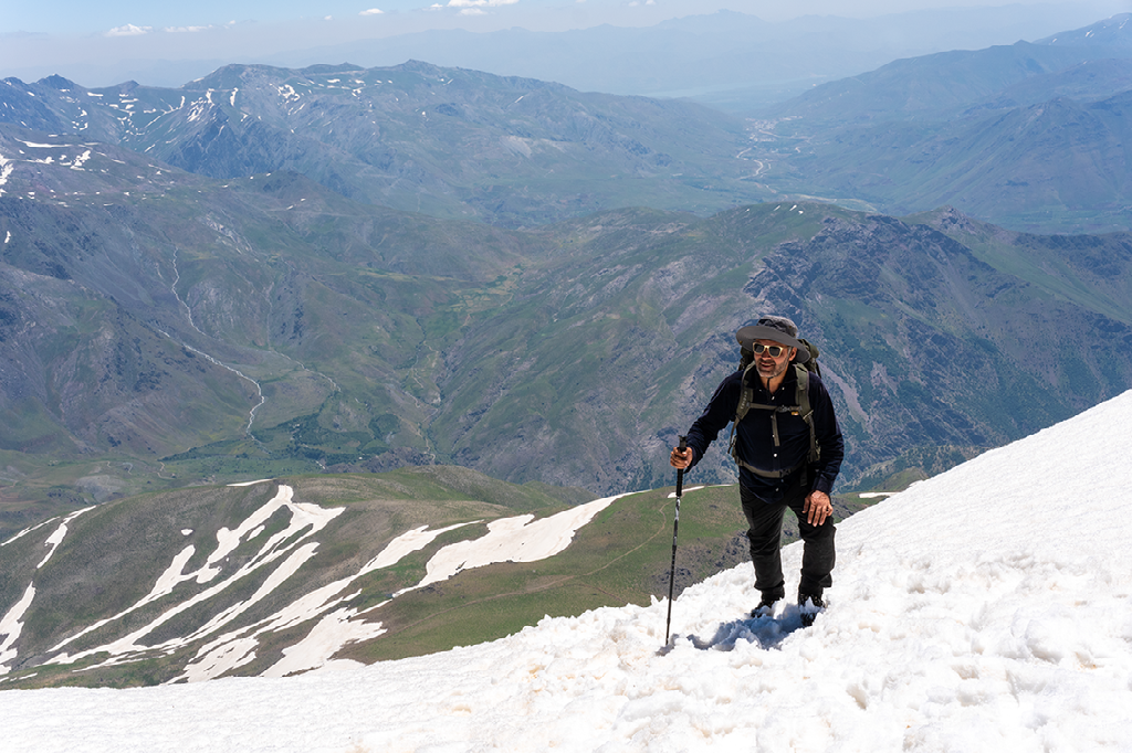 man met wandelstokken klimt door de sneeuw naar een bergtop met een uitgestrekt uitzicht over groene valleien en besneeuwde bergkammen