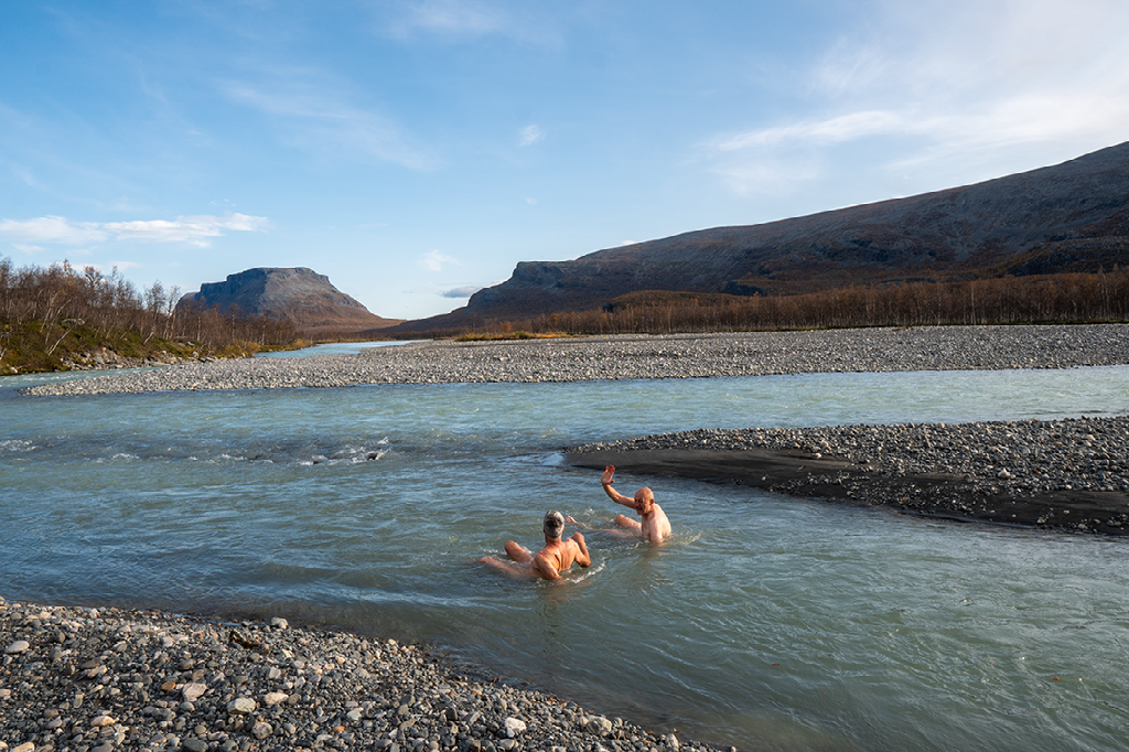 Twee mensen baden in een koude, ondiepe bergrivier omringd door grijze kiezels en met een kale berg op de achtergrond.