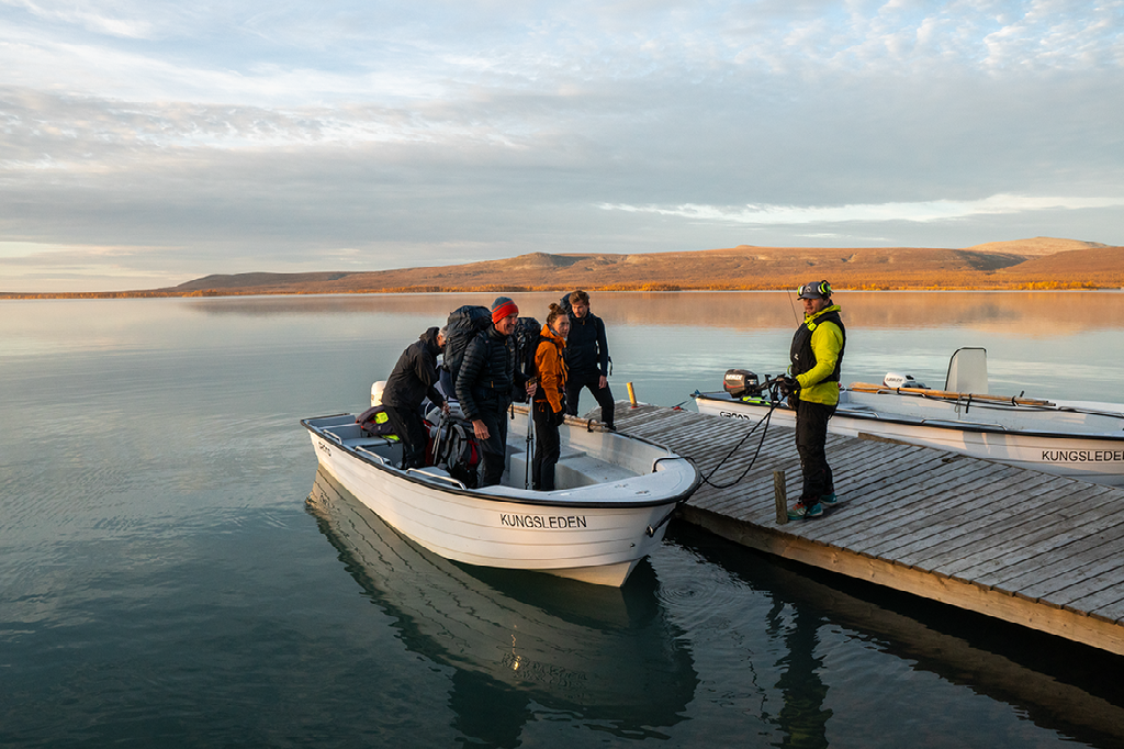 Een groep wandelaars stapt op een kleine witte boot aan een houten steiger aan een groot meer, met een lage horizon en lichte lucht.