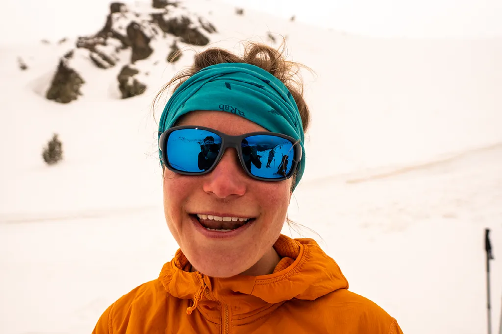 Close-up van een lachende vrouw met een turquoise hoofdband, blauwe zonnebril en een oranje jas in de sneeuw.