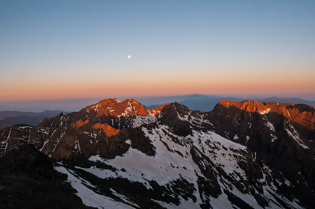 Bergtoppen, deels bedekt met sneeuw, worden verlicht door de laatste oranje zonnestralen tijdens zonsondergang of zonsopgang onder een heldere lucht met de maan.