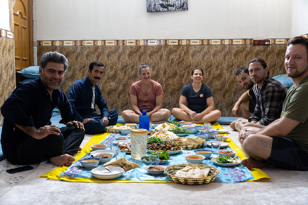 Een groep mensen zit in een kring op de grond rond een uitgebreide lokale maaltijd in een traditioneel huis
