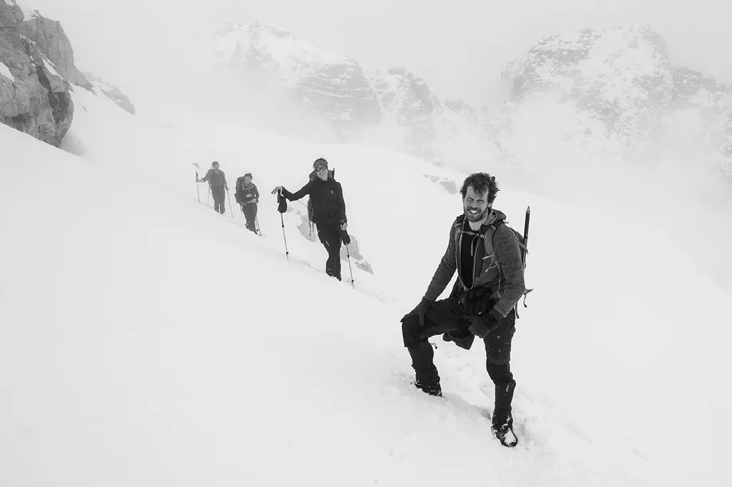 Een zwart-witfoto toont een lachende wandelaar met een rugzak en stokken die voorop loopt op een besneeuwde berghelling.
