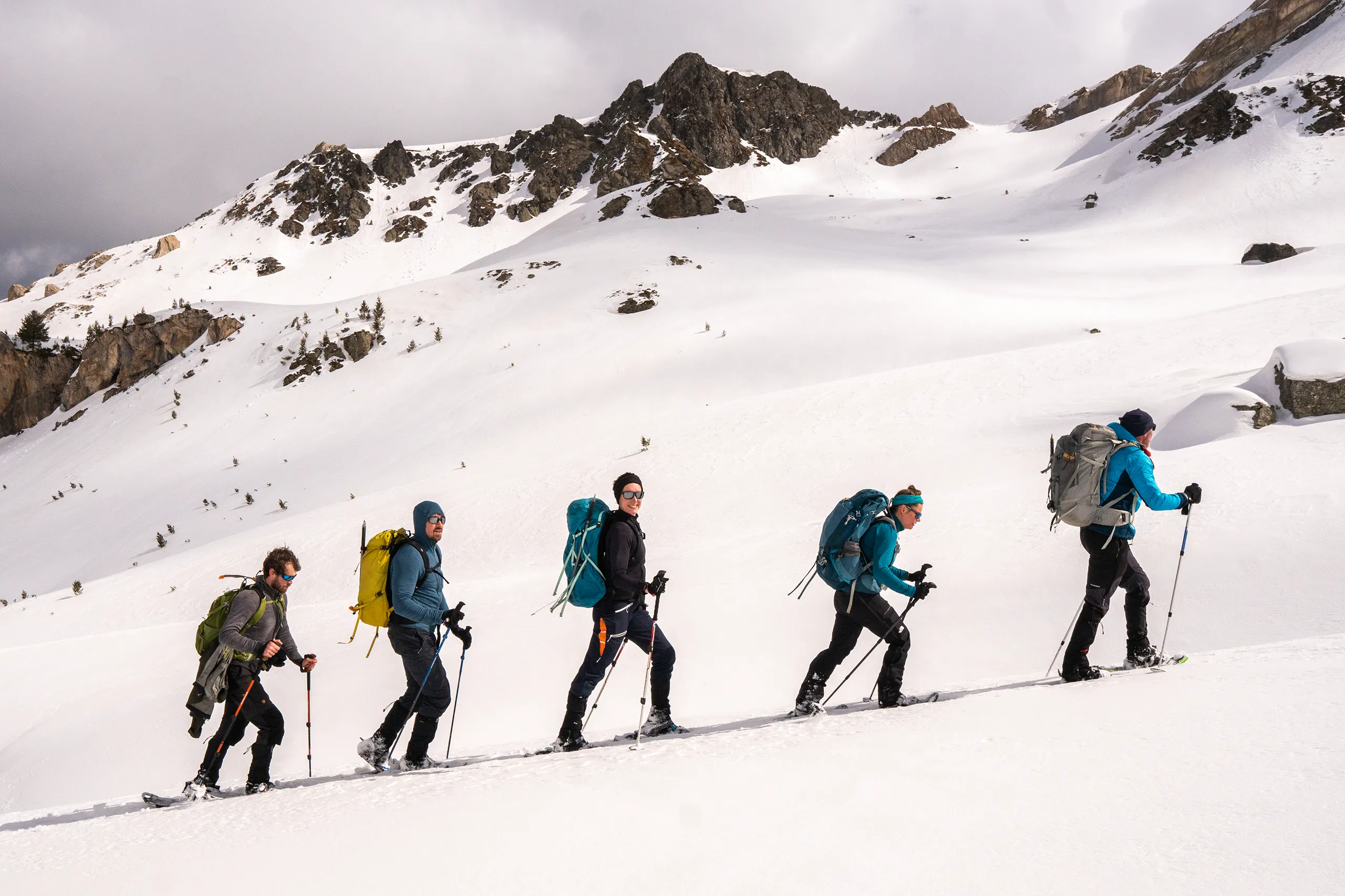 Vier wandelaars met rugzakken en stokken lopen achter elkaar een steile, besneeuwde berghelling op.