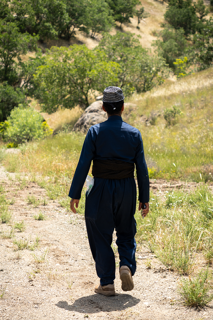Koerdische man in traditionele kleding wandelt over een stoffig bergpad tussen bomen en heuvels in het ruige landschap van Noord-Irak