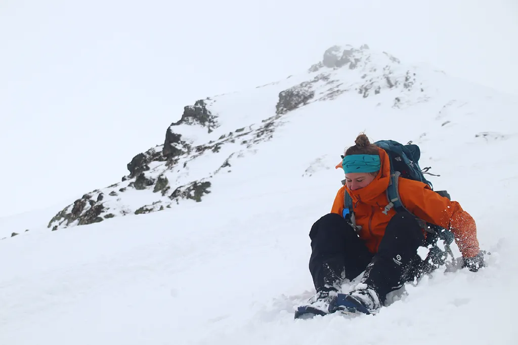 Een vrouwelijke wandelaar in een oranje jas glijdt of zit in de diepe sneeuw op een berghelling.