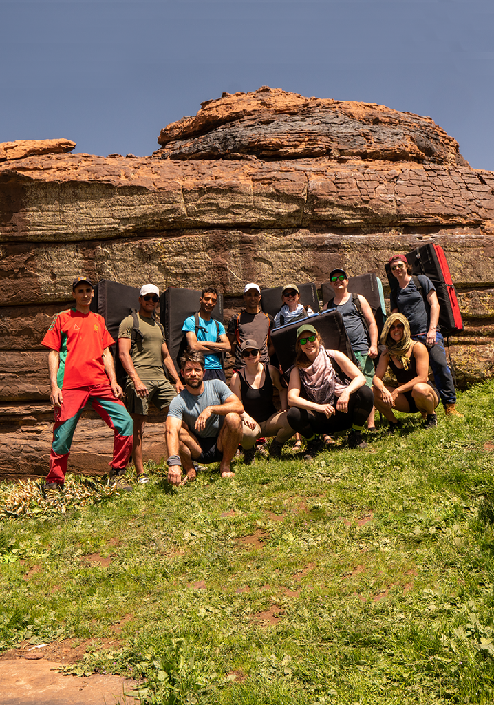 Een diverse groep mensen zit samen op een grasveld voor een grote, goudbruine rotsformatie onder een heldere, blauwe lucht.