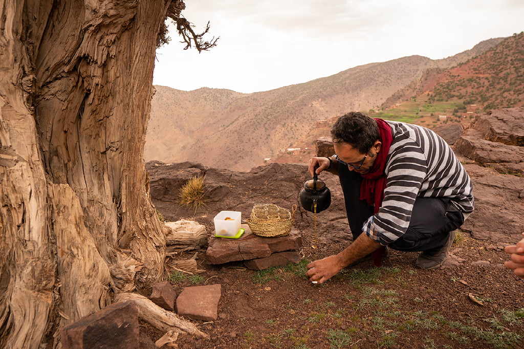 Een man in een gestreepte sjaal bereidt thee op een rotsachtige heuveltop met uitzicht op een droog berglandschap.