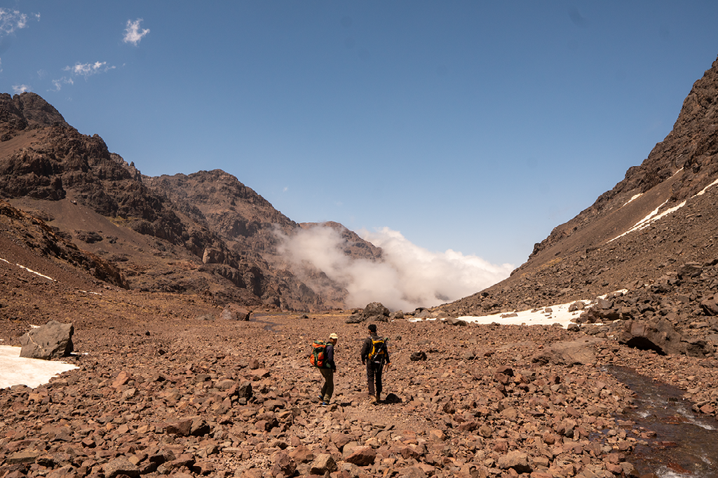 Twee wandelaars met rugzakken staan op een rotsachtige vlakte, kijkend naar de steile, bruine bergen en witte wolken.