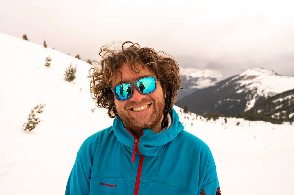 Portret van een lachende man met krullend haar, een blauwe jas en een blauwe zonnebril in een besneeuwde bergomgeving.