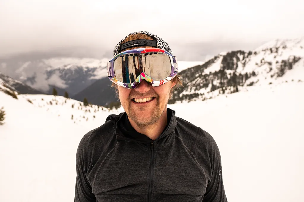 Close-up van een lachende man met een zwarte hoodie en een skibril op zijn hoofd in een besneeuwde omgeving.