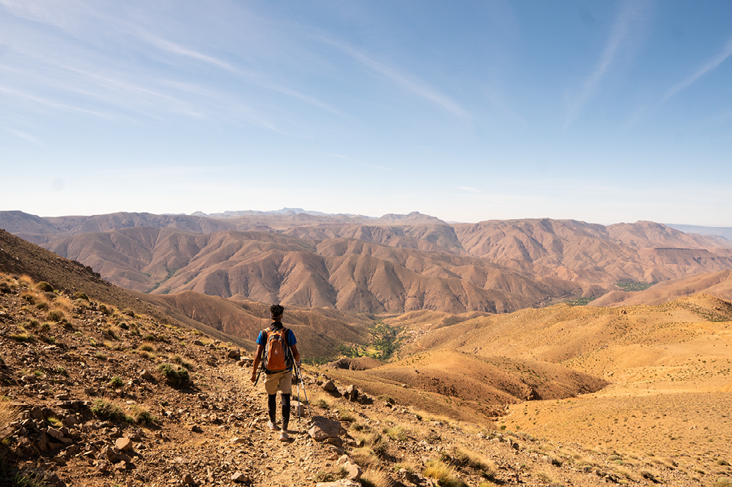 Een eenzame wandelaar met een rugzak loopt over een smal pad in een uitgestrekt, droog, goudkleurig berglandschap onder een helderblauwe lucht.