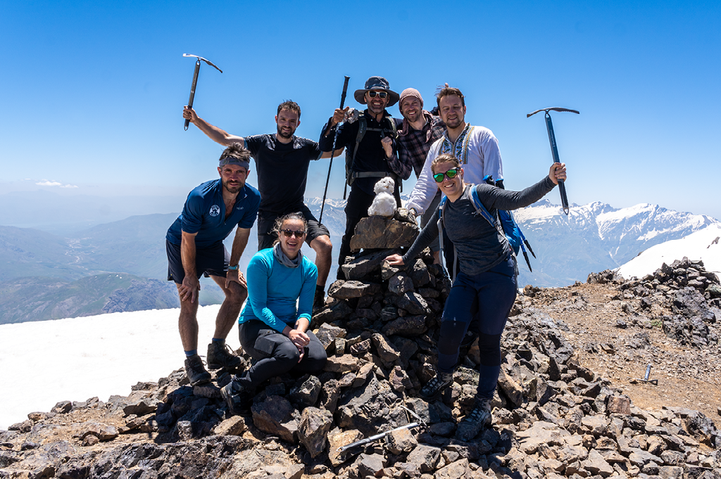 Groep bergbeklimmers poseert op de besneeuwde top van een berg in Koerdistan met uitzicht op omliggende bergketens en een helderblauwe lucht