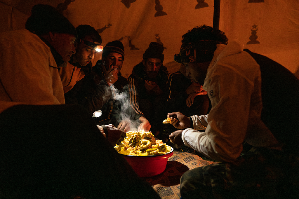 Een groep mannen zit in het donker rond een kom dampend voedsel, verlicht door een warme, oranje gloed in een tent of hut.