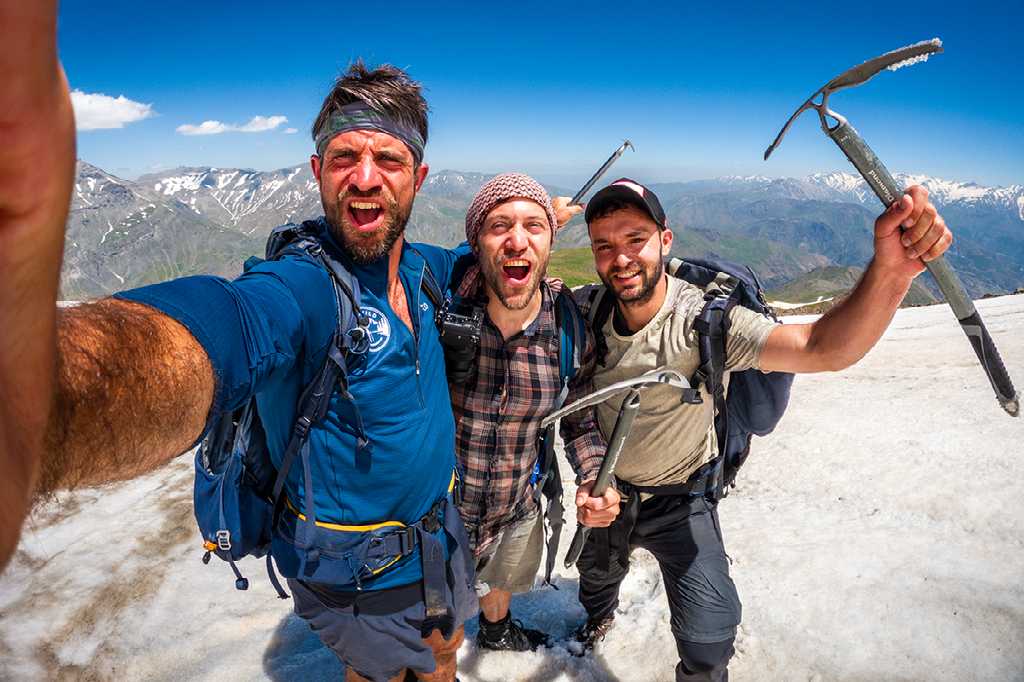 drie mannen maken een vrolijke selfie op een besneeuwde bergtop met ijsbijlen in de hand en uitzicht op een ruig berglandschap
