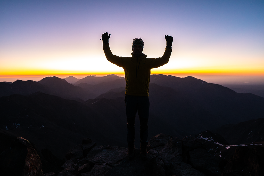 Een wandelaar staat op een bergtop met opgeheven armen, in silhouet tegen een gloeiende zonsondergang over een golvend berglandschap.