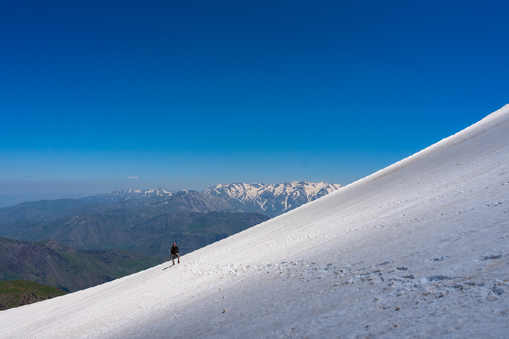 een bergbeklimmer loopt over een steile besneeuwde helling met een helderblauwe lucht en bergtoppen op de achtergrond