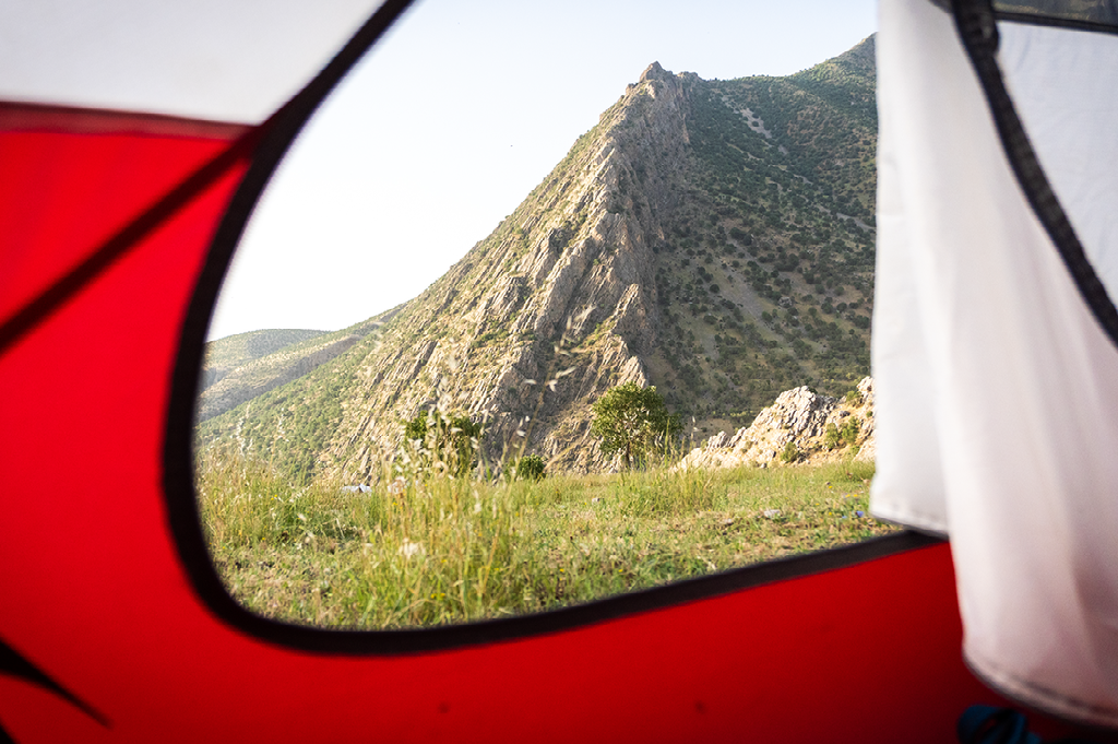 uitzicht op een ruige berghelling en groene vallei, geframed door de opening van een rood trekkingtentje in de ochtendzon