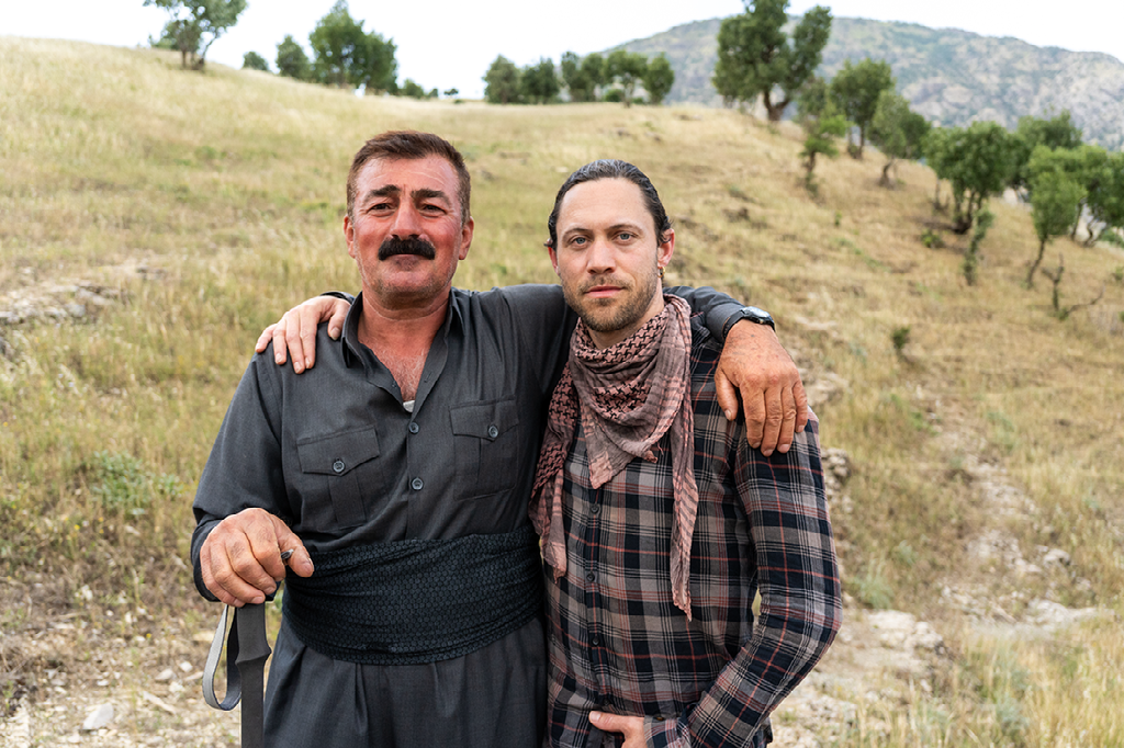 twee mannen poseren zij aan zij in een heuvelachtig landschap met gras en verspreide bomen, hun armen vriendschappelijk om elkaar heen