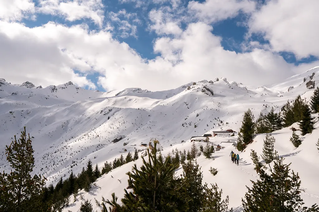 Twee wandelaars lopen op een besneeuwde heuvel, met dennenbomen, een paar hutten en besneeuwde bergen op de achtergrond.