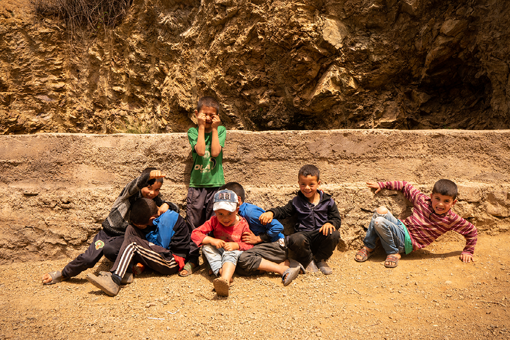 Vijf lachende Berberkinderen in casual kleding zitten op een zandvlakte voor een rotswand.