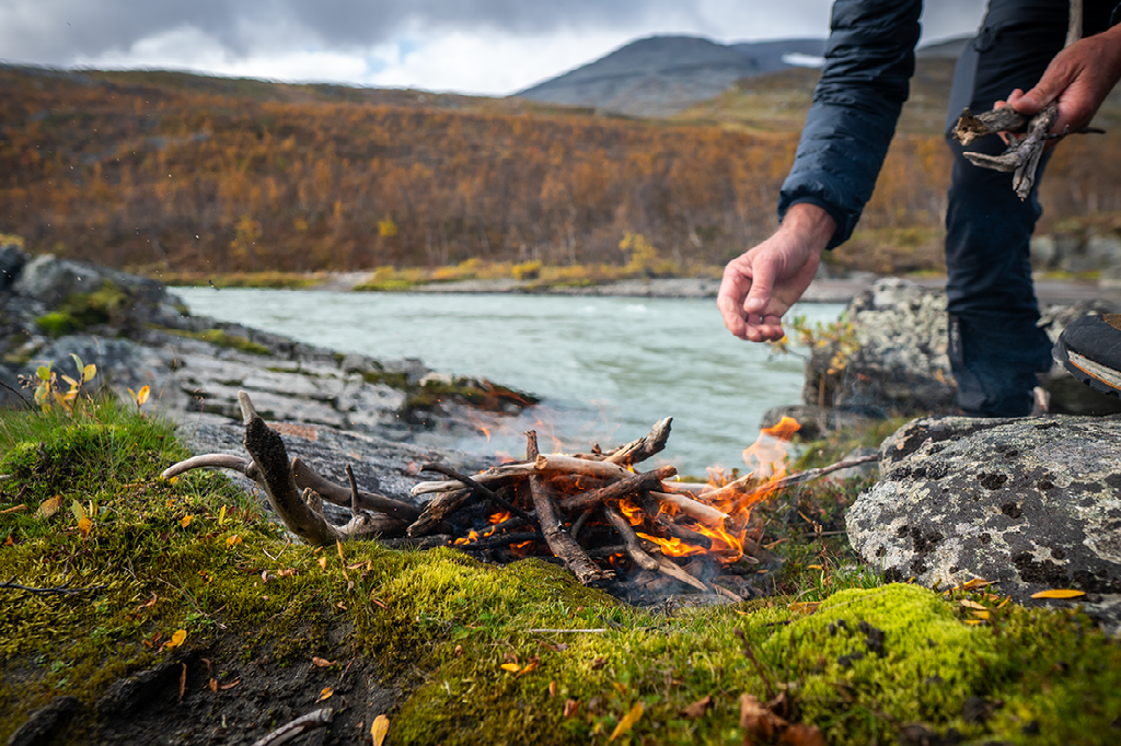 Iemand steekt een klein kampvuur aan op de rotsachtige, mosbegroeide oever van een rivier in een berglandschap met herfstkleuren.
