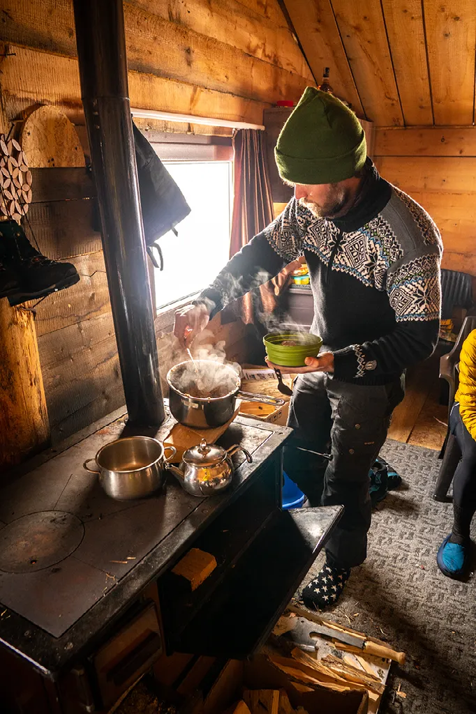 Een man in een muts en een trui kookt in metalen potten op een houtkachel in een houten hut.