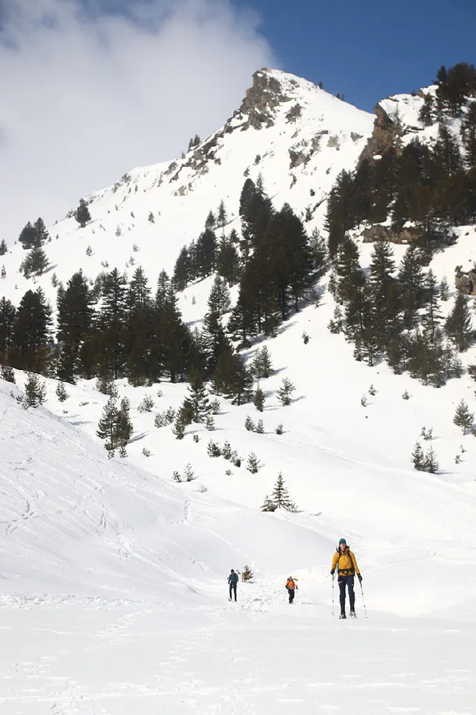 Drie wandelaars, waaronder één met een gele jas, klimmen een besneeuwde helling op richting besneeuwde bergen en dennenbomen.