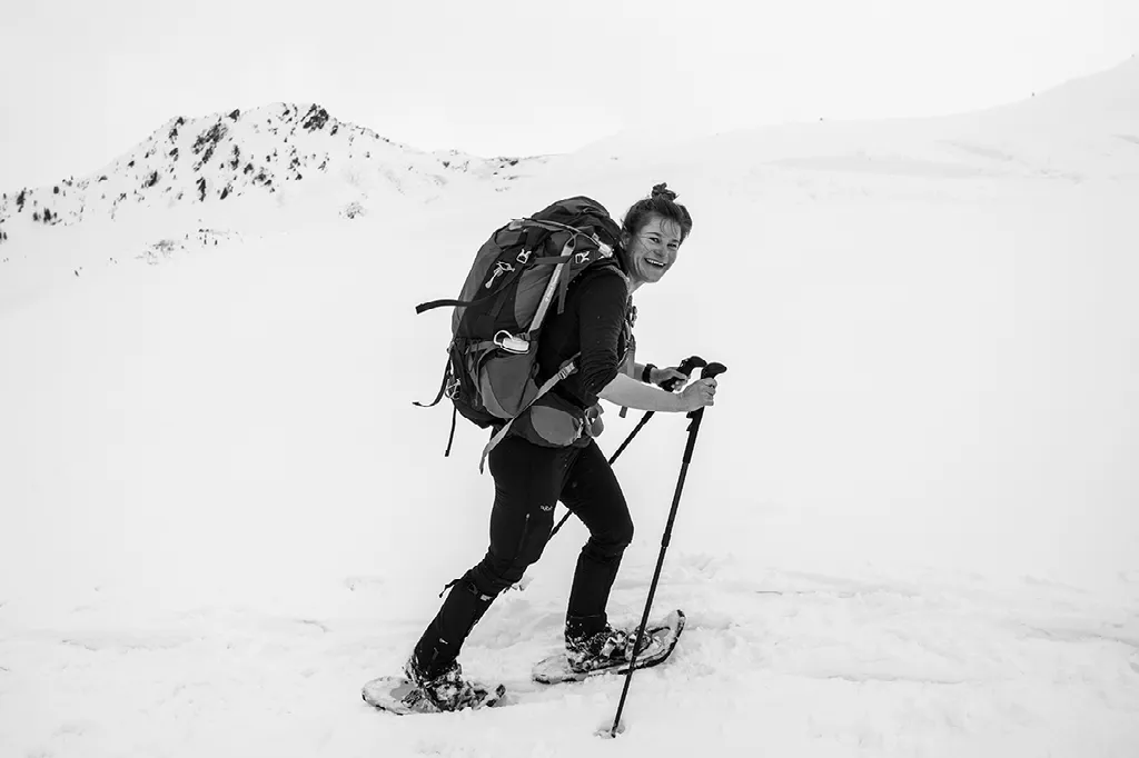 Een zwart-witfoto van een lachende vrouwelijke wandelaar met een grote rugzak, stokken en sneeuwschoenen in een besneeuwde omgeving.