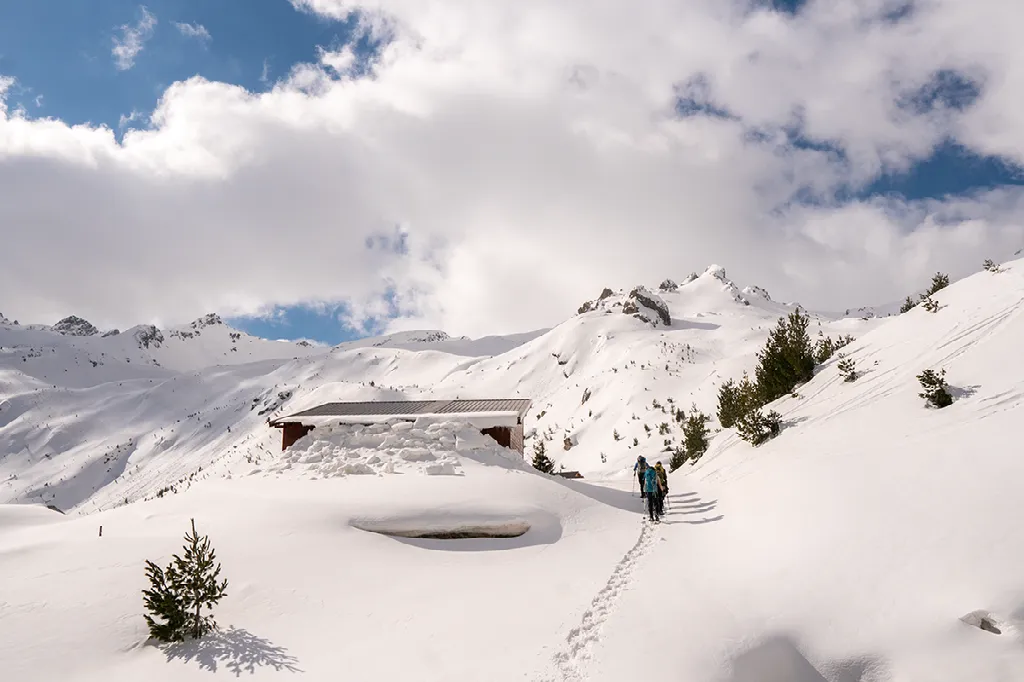 Een wandelaar loopt door diepe sneeuw in de richting van een hut, met besneeuwde bergen op de achtergrond.