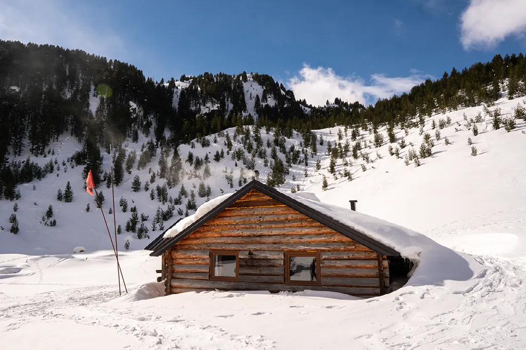 Een met sneeuw bedekte houten hut staat op een besneeuwde helling met een dicht bebost berglandschap op de achtergrond.