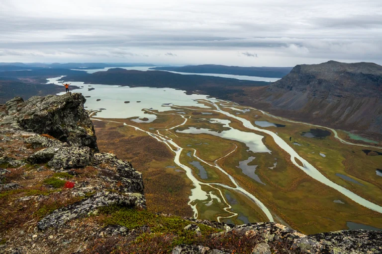 Een klein figuur staat op een rotsachtige klif met een weids uitzicht over de kronkelende rivierdelta en meren in de Rapa-vallei in Sarek Nationaal Park.