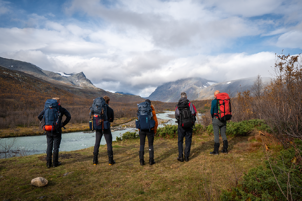 Vier wandelaars met grote rugzakken kijken naar een bergbeek en een ruig gebergte, staand in een herfstachtig landschap.