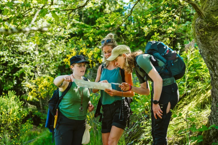 Drie wandelaars, twee met rugzakken, staan in een bos en kijken naar een kaart of mobiele telefoon.