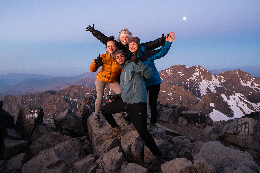 Vier enthousiaste wandelaars juichen op een rotsachtige bergtop bij zonsondergang, met besneeuwde bergen op de achtergrond.
