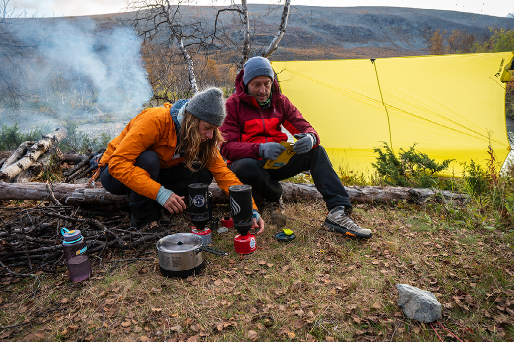 Twee wandelaars koken een maaltijd op een draagbare brander buiten hun felgele tent in een bosrijke, herfstachtige omgeving.